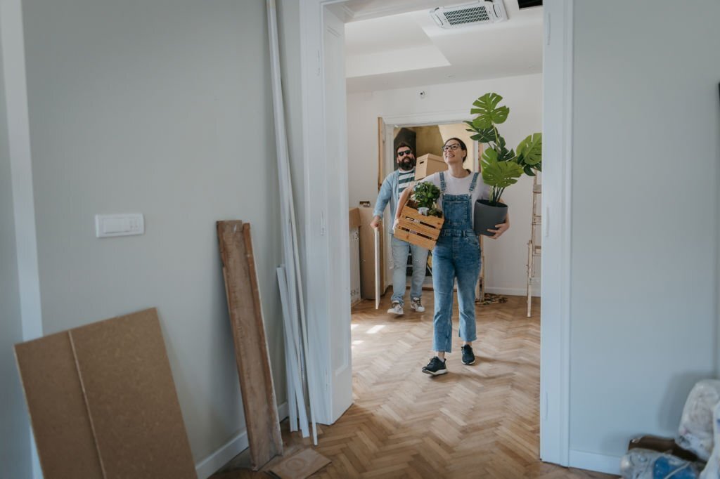 Couple moving in their new apartment, woman is carrying potted plant and wooden box.