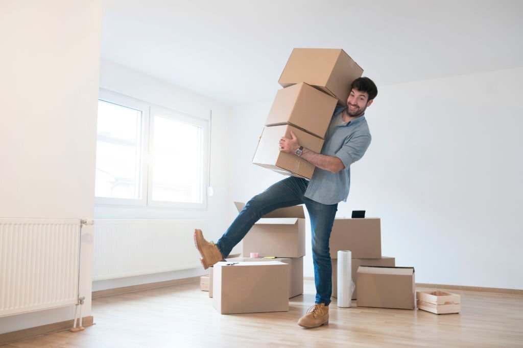 Funny young man clumsy carrying a stack of cardboard boxes while moving into a new apartment.