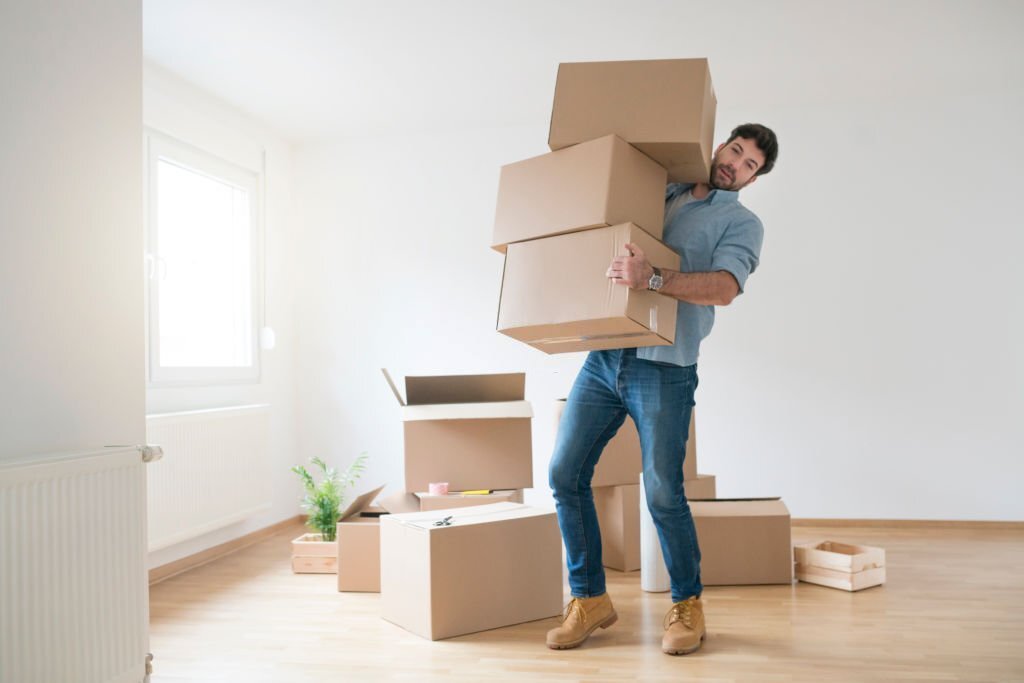 Funny young man clumsy carrying a stack of cardboard boxes while moving into a new apartment.