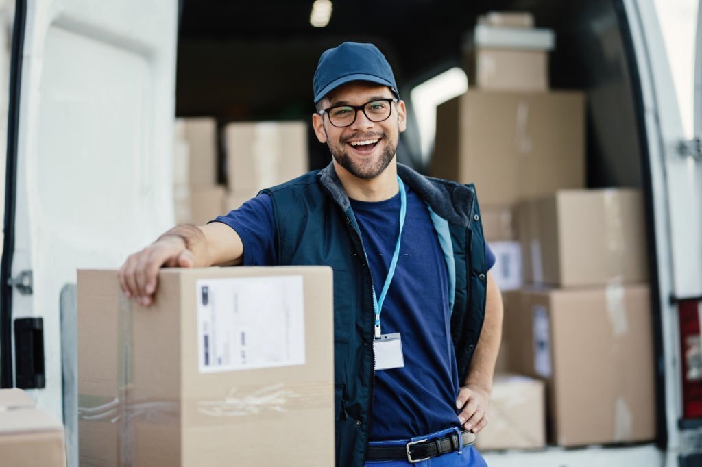 Young happy delivery man loading cardboard boxes in a van and looking at camera.